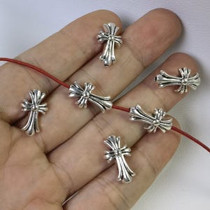 May include: A close-up of a silver cross charm necklace with a red cord. The cross charms are detailed and have a slightly textured finish.