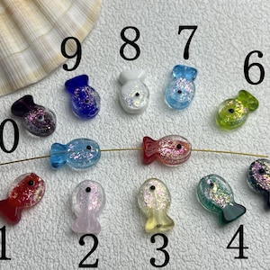 Może przedstawiać: Kolekcja kolorowych, szklanych koralików w kształcie ryb z brokatowymi akcentami. Koraliki są w różnych kolorach, w tym czerwonym, niebieskim, zielonym i fioletowym. Są nawleczone na cienki złoty drucik, idealne do tworzenia biżuterii.