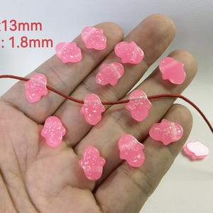 Peut inclure: Un ensemble de perles en forme de main de Hamsa roses de 13 mm avec un trou de 1,8 mm. Les perles sont enfilées sur un cordon rouge.