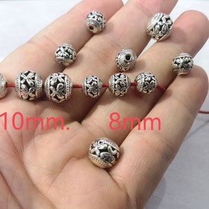 May include: A collection of round, silver-toned metal beads featuring an embossed floral pattern and dark recessed detailing. The beads are shown in two sizes, labeled as 10mm and 8mm, with some beads strung on a thin red cord and others loose.