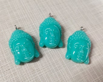 May include: Three turquoise Buddha head pendants with silver-colored loops for hanging. The pendants are made of a glossy material and feature detailed facial features and a serene expression. The heads are in various positions.