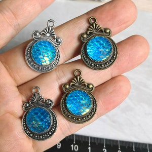 May include: Four silver and bronze pendant charms with blue iridescent mermaid scale designs. Each charm features a detailed crown-like design.