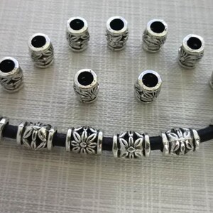 K&ouml;nnte beinhalten: Eine silberne Halskette mit einem schwarzen Band und silbernen Perlen mit floralem Muster. Es befinden sich 12 lose Perlen auf dem Bild.
