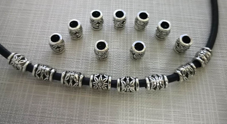 K&ouml;nnte beinhalten: Eine silberne Halskette mit einem schwarzen Band und silbernen Perlen mit floralem Muster. Es befinden sich 12 lose Perlen auf dem Bild.