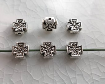 May include: Six silver-toned metal beads shaped like crosses with an embossed floral pattern in the centre. Three of the beads are arranged in a row on a thin green wire, while the other three are positioned above them in a horizontal line.