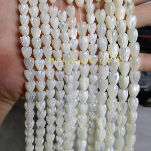 May include: Multiple strands of white mother of pearl beads carved into small tulip or flower shapes. Each bead features a smooth, iridescent finish with vertical ridges. The strands are arranged vertically, showcasing the uniform size and shape of the individual shell components.