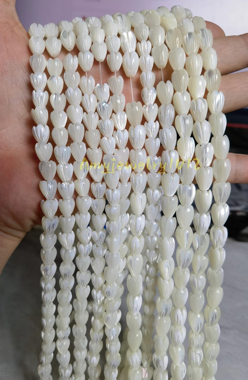 May include: Multiple strands of white mother of pearl beads carved into small tulip or flower shapes. Each bead features a smooth, iridescent finish with vertical ridges. The strands are arranged vertically, showcasing the uniform size and shape of the individual shell components.