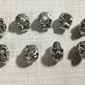 Peut inclure: Huit perles en argent en forme de crâne avec un trou en haut. Les perles sont toutes de designs différents et ont un aspect légèrement vieilli.
