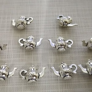 Könnte beinhalten: Eine Sammlung kleiner, silberfarbener Teekannen-Anhänger. Jeder Anhänger hat ein detailliertes Design, einschließlich Ausguss, Henkel und Deckel. Einige Teekannen tragen das Wort "time". Ideal für die Schmuckherstellung oder zum Basteln.