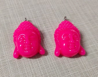 May include: Two bright pink Buddha head pendants. Each pendant features a serene face with closed eyes and a small loop at the top for hanging. The pendants are made of a glossy material.