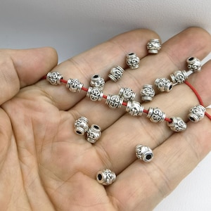 May include: A collection of small, barrel-shaped silver-toned metal beads featuring an intricate embossed scrollwork pattern. Several beads are strung onto a thin red cord, while others are scattered loosely, showcasing their cylindrical shape.