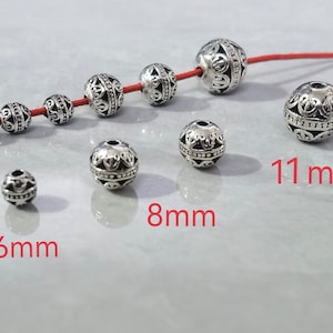 May include: A collection of round, silver-toned metal beads with intricate embossed patterns. Some beads are strung on a red cord, while others are loose. The image displays three distinct sizes labeled as 6mm, 8mm, and 11mm in red text.