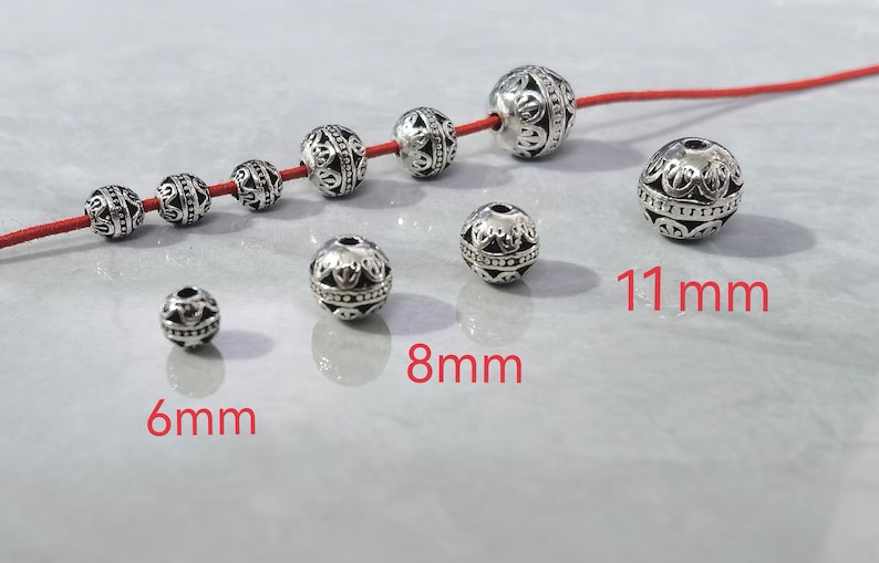 May include: A collection of round, silver-toned metal beads with intricate embossed patterns. Some beads are strung on a red cord, while others are loose. The image displays three distinct sizes labeled as 6mm, 8mm, and 11mm in red text.