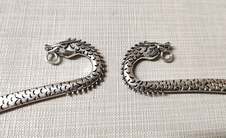 May include: Two silver-toned metal bookmarks, each featuring a detailed dragon head at the curved top. The dragon design includes textured scales along the neck and body, with a small circular loop attached beneath the chin for hanging charms.