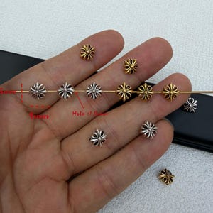 May include: A collection of small, cross-shaped beads in silver and gold tones. The beads measure 9mm in diameter with a 1.5mm hole. Several beads are threaded onto a thin metal rod, while others are scattered, showcasing the detailed design.