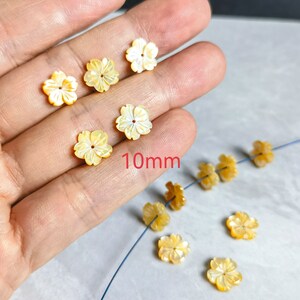 May include: A strand of 10mm shell flower beads in a pale yellow colour. The beads are strung on a thin wire and are arranged in a line. There are also several loose beads in the image.