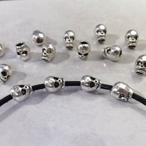 May include: A collection of small, silver-colored metal skull beads. Several beads are scattered loosely, while four identical skull beads are threaded onto a single, thin black leather cord that curves across the bottom of the frame.