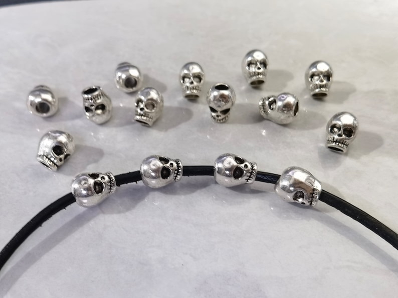May include: A collection of small, silver-colored metal skull beads. Several beads are scattered loosely, while four identical skull beads are threaded onto a single, thin black leather cord that curves across the bottom of the frame.