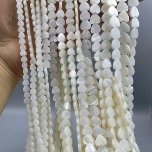 Könnte beinhalten: Eine Strähne aus weißen Muschelperlen in Herzform. Die Perlen sind auf einem durchsichtigen Faden aufgefädelt und haben einen Durchmesser von etwa 1,2 cm.