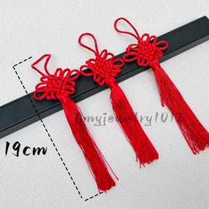 Può includere: Tre nappe decorative rosso brillante con nodi complessi sulla parte superiore e anelli per appenderle. Ogni nappa misura 19 cm di lunghezza totale. Il set è disposto in fila, caratterizzato da un colore rosso vivace e frange lunghe.