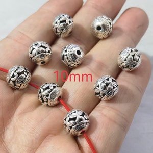 May include: A collection of eight spherical silver-toned beads, each featuring an intricate embossed floral pattern and a central hole. The beads measure 10mm in diameter. Some beads are strung on a thin red cord, while others are loose.