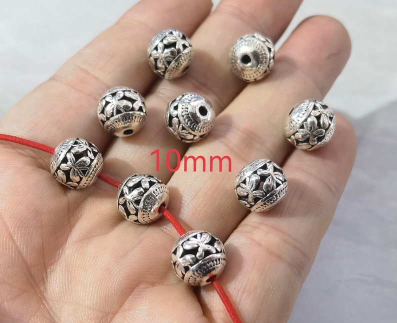 May include: A collection of eight spherical silver-toned beads, each featuring an intricate embossed floral pattern and a central hole. The beads measure 10mm in diameter. Some beads are strung on a thin red cord, while others are loose.