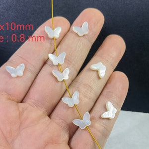 May include: Small, white butterfly-shaped beads with a pearlescent sheen. The beads are 7mm x 10mm with a 0.8 mm hole. Several beads are strung on a thin, yellow thread. The beads are displayed against a dark background.