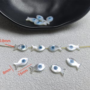 Może przedstawiać: Białe koraliki w kształcie rybki z niebieskimi detalami oczu, o wymiarach 15 mm długości i 8 mm szerokości. Koraliki są nawleczone na cienki złoty drucik. Kilka koralików znajduje się w czarnej miseczce. Otwór ma 0,8 mm.