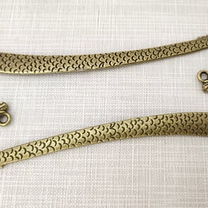 May include: Two antique brass-colored metal bookmarks, each featuring a long, flat body with a textured scale pattern and a curved end shaped like a dragon head with a small circular loop attached near the top of the dragon's head.