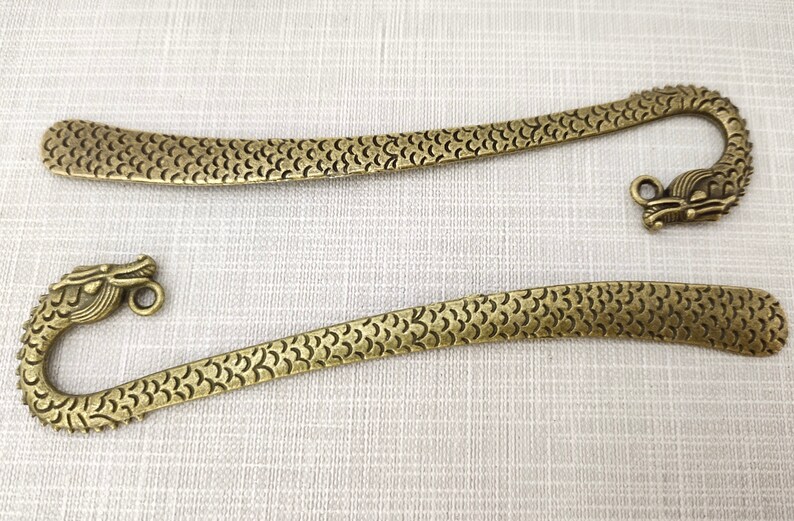 May include: Two antique brass-colored metal bookmarks, each featuring a long, flat body with a textured scale pattern and a curved end shaped like a dragon head with a small circular loop attached near the top of the dragon's head.