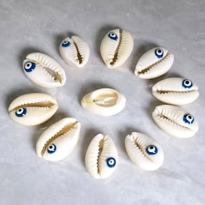 Könnte beinhalten: Zehn weiße Muscheln mit blauen bösen Augen-Anhängern. Die Muscheln sind in einem Kreis auf einer hellgrauen Oberfläche angeordnet.
