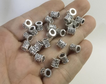Pode incluir: Uma coleção de pequenas contas cilíndricas de metal com um acabamento prateado brilhante e um intrincado padrão floral ou de volutas em relevo que cobre a superfície exterior. Cada conta apresenta um orifício central para enfiar.