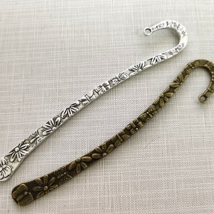 Può includere: Due segnalibri in metallo, uno color argento e uno color bronzo antico. Entrambi presentano un motivo in rilievo di fiori e farfalle lungo tutta la lunghezza, terminando con un gancio ricurvo dotato di un piccolo occhiello circolare.
