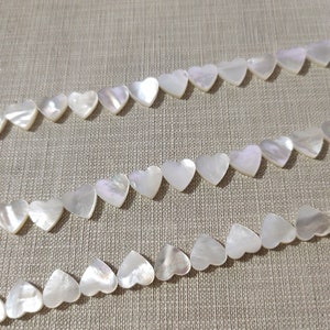 May include: A strand of white mother of pearl heart-shaped beads. The beads are strung on a white thread and are arranged in a row.