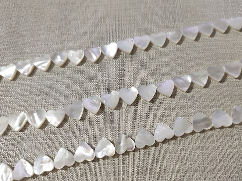 May include: A strand of white mother of pearl heart-shaped beads. The beads are strung on a white thread and are arranged in a row.