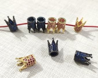 May include: A collection of crown-shaped beads featuring metallic finishes in black and gold. The black beads are encrusted with blue stones, while the gold beads are encrusted with pink stones. Some beads are threaded on a thin red cord.
