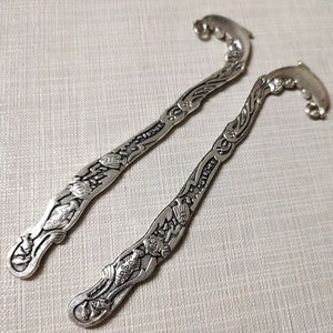 May include: Two silver bookmark pendants with intricate designs. The bookmarks are shaped like a curved line with a decorative end. The designs feature a variety of patterns and symbols.