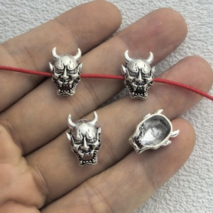 May include: Silver-colored metal beads shaped like Japanese Oni masks, with horns and detailed facial features. The beads are strung on a red cord, with one bead detached. The beads are small, and the details are intricate.