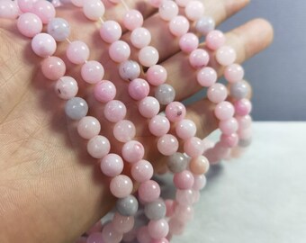 Puede incluir: Se muestran varias hebras de cuentas de piedras preciosas rosas, cada una de aproximadamente 8 mm de diámetro. Las cuentas varían en tonos de rosa y gris, ensartadas para formar collares o pulseras. La imagen muestra la superficie lisa y pulida de las cuentas.