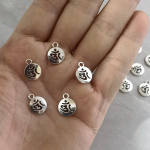 May include: A collection of silver-toned metal charms featuring the Om symbol. The charms are round and have a hole at the top for attaching to jewelry or other crafts.