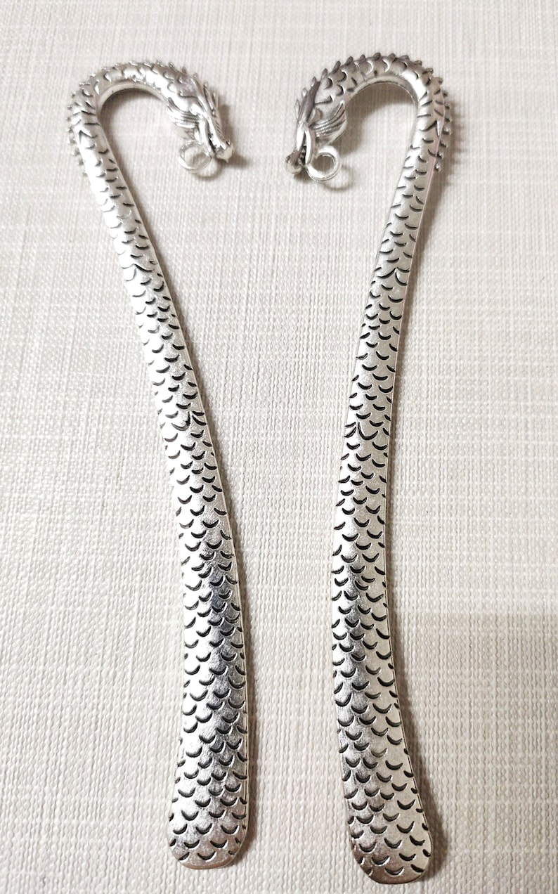 May include: Two silver-toned metal bookmarks, each featuring a curved top shaped like a dragon head with detailed scales along the length of the body. The dragon heads are oriented toward each other, and each includes a small circular loop at the chin.