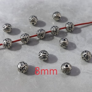 May include: A collection of spherical silver-toned metal beads, each featuring an intricate embossed floral pattern. Four beads are threaded onto a thin red cord, while others are scattered nearby. The text 8mm is visible in red.