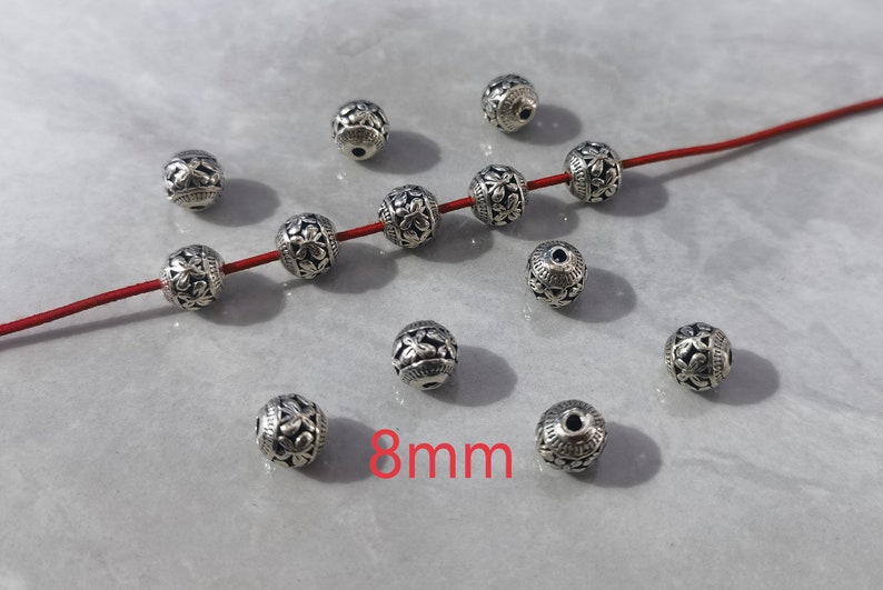 May include: A collection of spherical silver-toned metal beads, each featuring an intricate embossed floral pattern. Four beads are threaded onto a thin red cord, while others are scattered nearby. The text 8mm is visible in red.