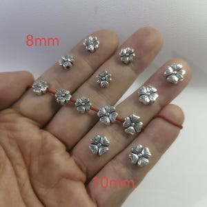 May include: Silver flower-shaped metal beads in two sizes, 8mm and 10mm. The beads are arranged on a hand, with the smaller beads on the left and the larger beads on the right.
