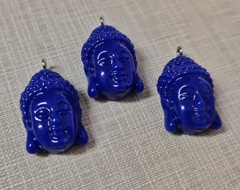 May include: Three blue resin Buddha head pendants with silver jump rings. The pendants are detailed with a textured hair design and a serene facial expression.