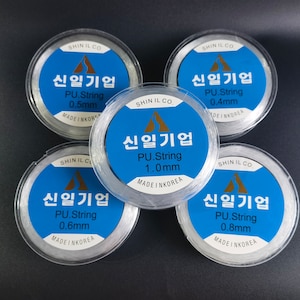 May include: Five spools of clear elastic string, each with a label in Korean that reads "Shin Il Gi Up" and indicates the string's thickness in millimeters. The spools are arranged in a circle on a black surface.