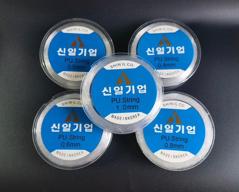 May include: Five spools of clear elastic string, each with a label in Korean that reads "Shin Il Gi Up" and indicates the string's thickness in millimeters. The spools are arranged in a circle on a black surface.