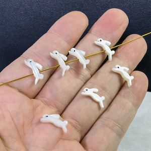 May include: Seven white porcelain bunny figurines, each with a different pose, are arranged on a hand. The figurines are strung together with a gold wire.
