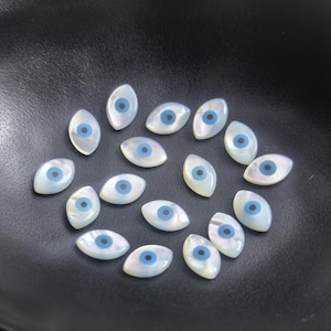 May include: A collection of small, oval-shaped, mother-of-pearl beads with a blue eye design. The beads are arranged on a dark, textured surface. These beads are ideal for jewelry making and craft projects.