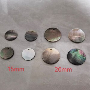 May include: Eight round, iridescent, black mother of pearl shell discs with a hole in the center. The discs are arranged in two rows of four. The top row has four 15mm discs and the bottom row has four 20mm discs. The discs are on a white surface.